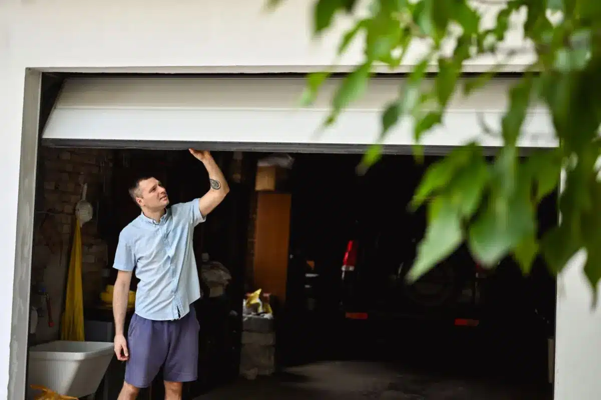 Man standing in a garage, lifting the door with one hand while looking up at it.